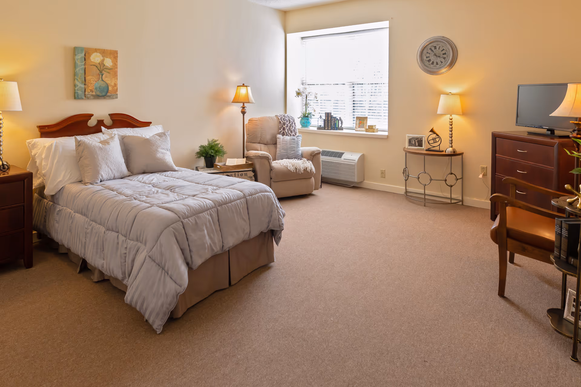 A cozy bedroom with a neatly made bed featuring a gray comforter and multiple pillows. There are two bedside tables with lamps on each side of the bed. A comfortable armchair with pillows and a throw blanket is placed near a window with blinds. The room also includes a wooden dresser with a TV on top, a round side table with decorative items, a wall clock, and a wooden chair. The walls are light-colored, and the floor is carpeted.