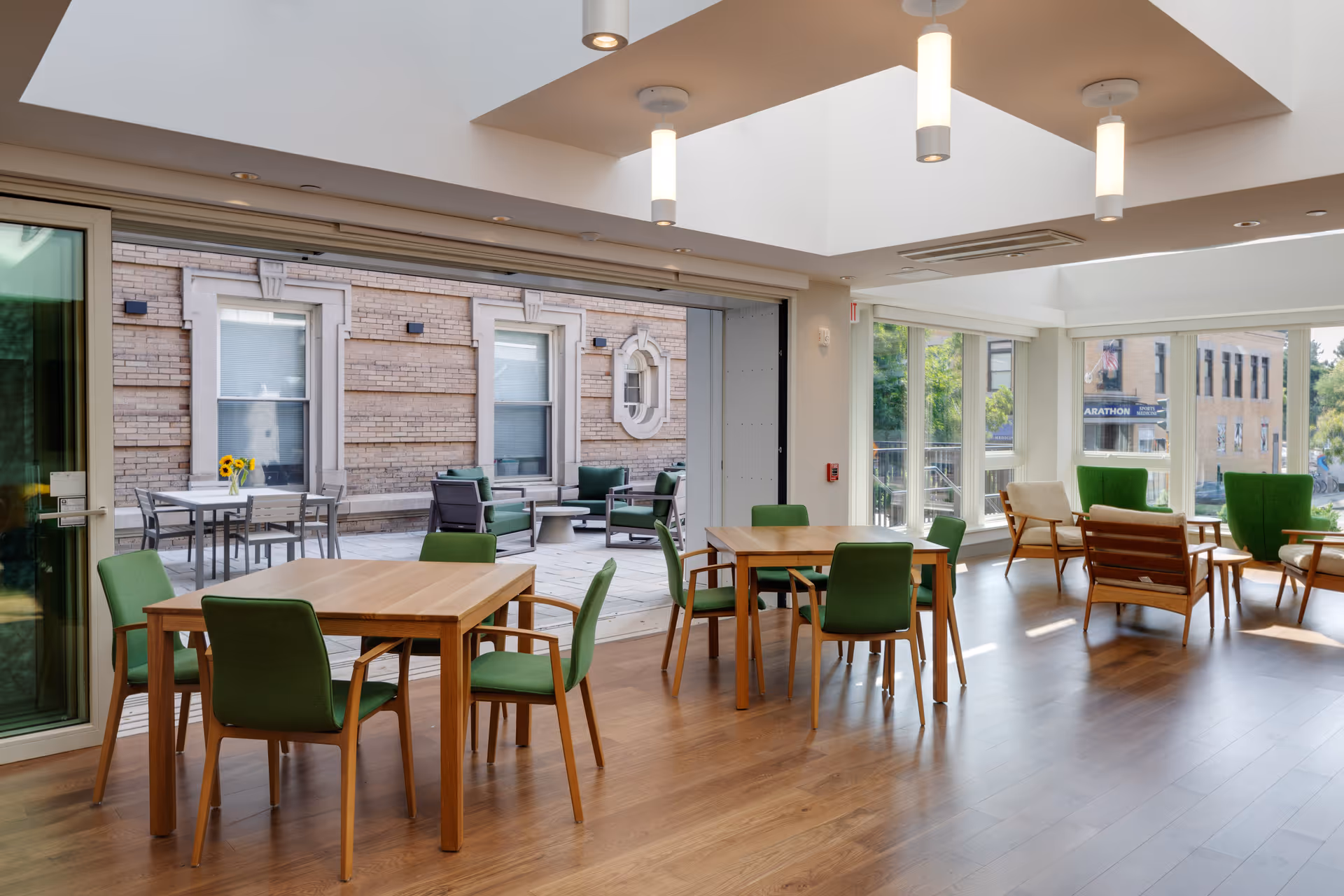 Bright communal dining and lounge area with wooden tables and green chairs opening onto an outdoor patio through large sliding glass doors.
