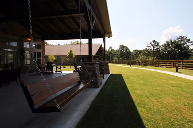A covered outdoor patio area with a wooden swing hanging from the ceiling by metal chains. The patio has stone pillars and several chairs and tables. Beyond the patio is a well-maintained grassy lawn with a paved walkway, wooden fence, and trees in the background under a clear sky.