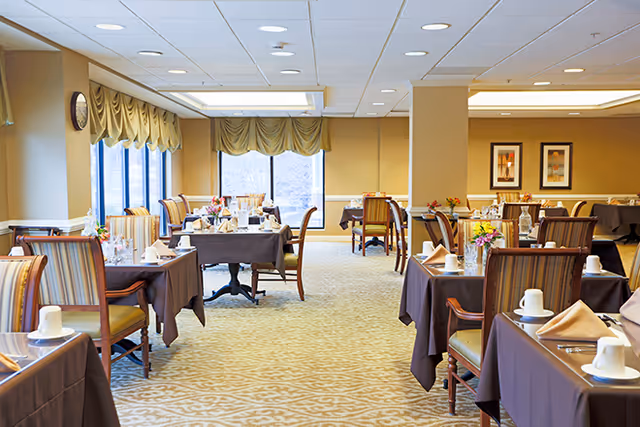 Spacious dining room with neatly set tables covered in brown tablecloths, upholstered chairs, and large windows.