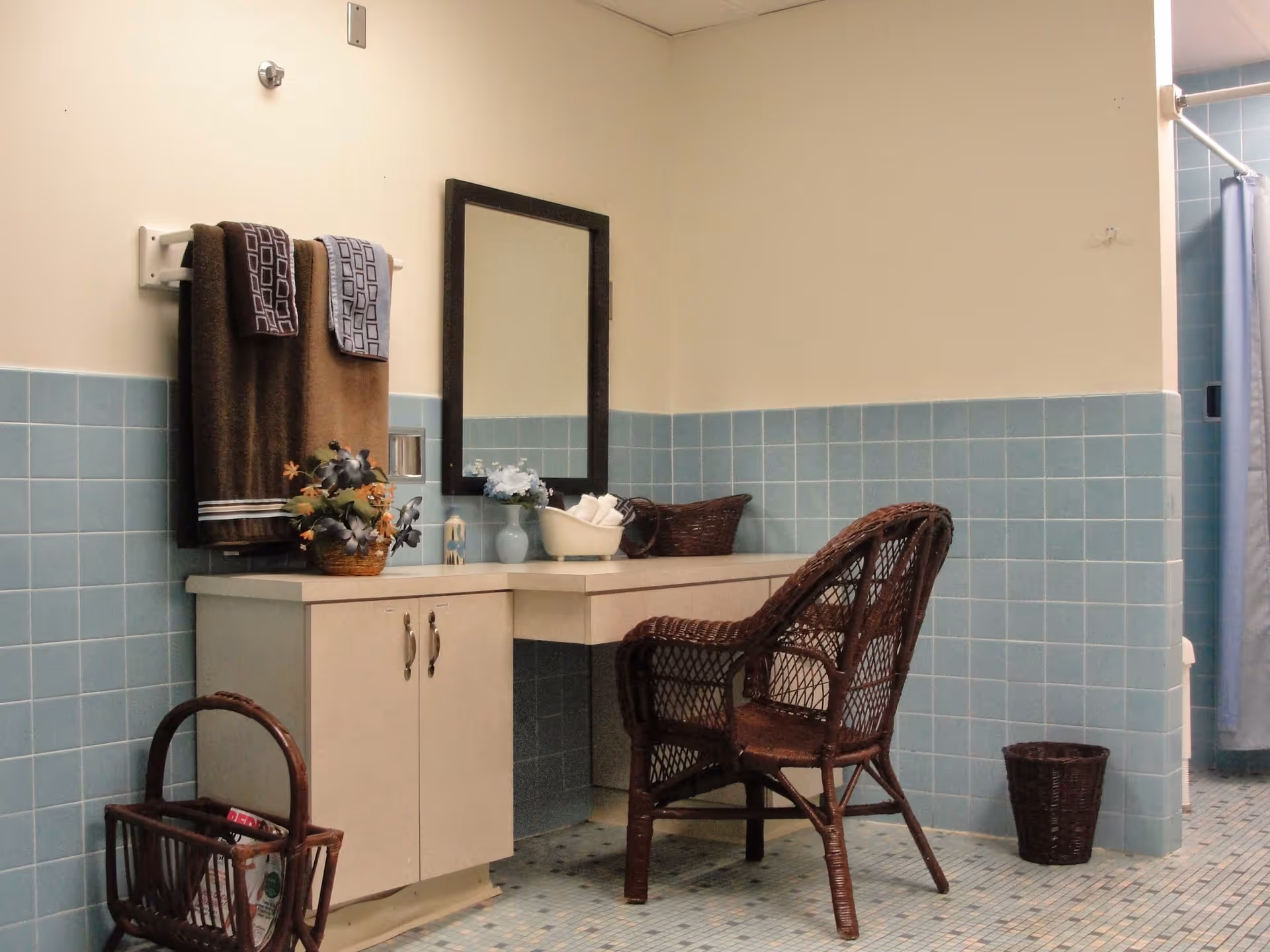 A tiled bathroom vanity area with a mirror, wicker chair, towels and decorative baskets.