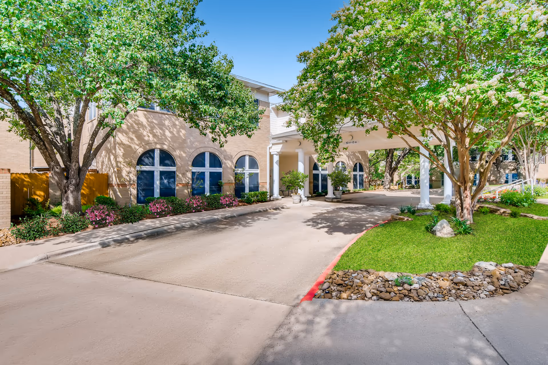 Entrance area of Asher Point Senior Living of Austin showing a driveway with a covered drop-off area supported by white columns, surrounded by green trees, shrubs, and landscaped grass with rocks.
