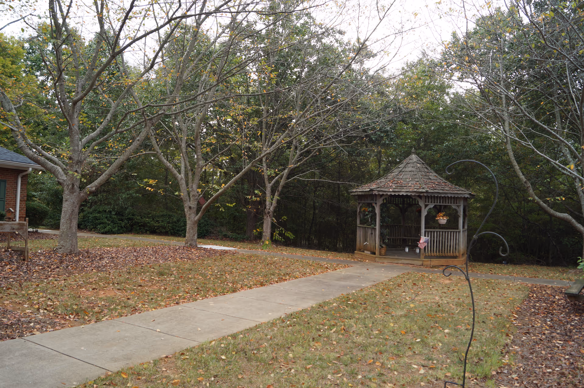 A peaceful outdoor area with a concrete walkway leading to a wooden gazebo surrounded by trees with some fallen leaves on the grass and ground, suggesting an autumn setting.