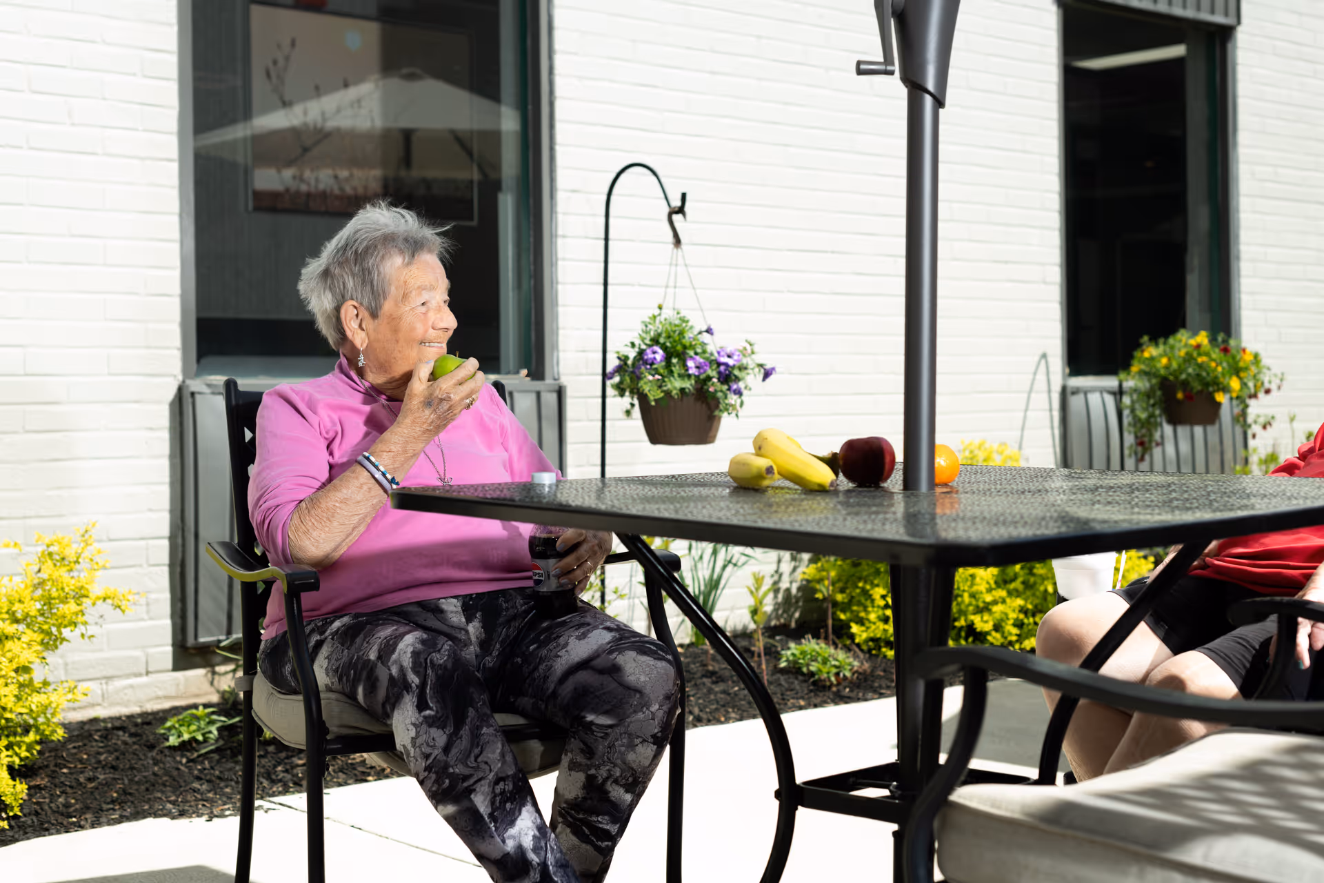 An elderly woman wearing a pink long-sleeve shirt and patterned pants sits outdoors at a metal table with a black umbrella pole in the center. She is holding a green apple and smiling, with a small container in her other hand. On the table are bananas, an apple, and an orange. Behind her is a white brick building with windows and hanging flower pots, along with some yellow and green plants in the garden bed.
