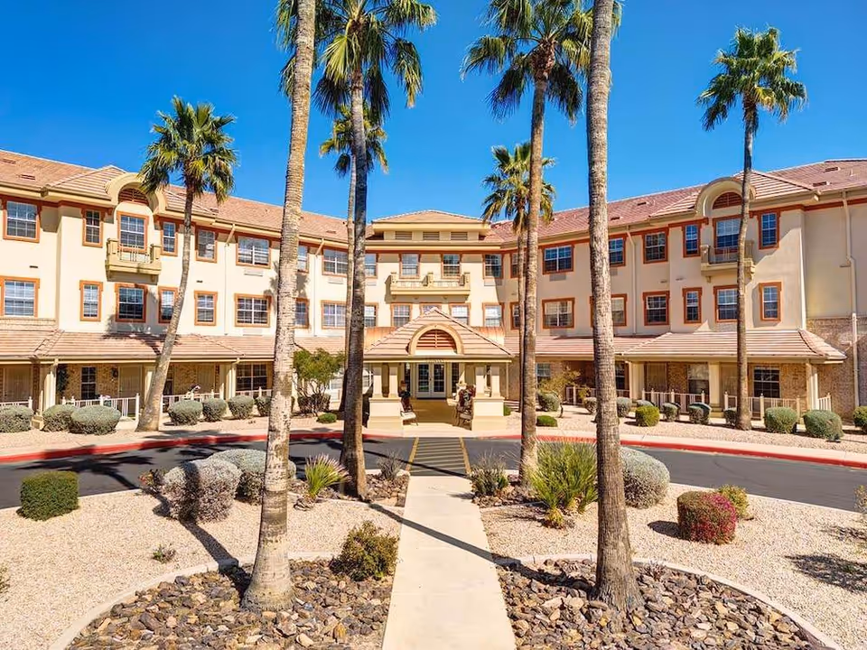 Exterior view of a three-story assisted living facility with beige walls and red-tiled roof, surrounded by palm trees and desert landscaping under a clear blue sky.