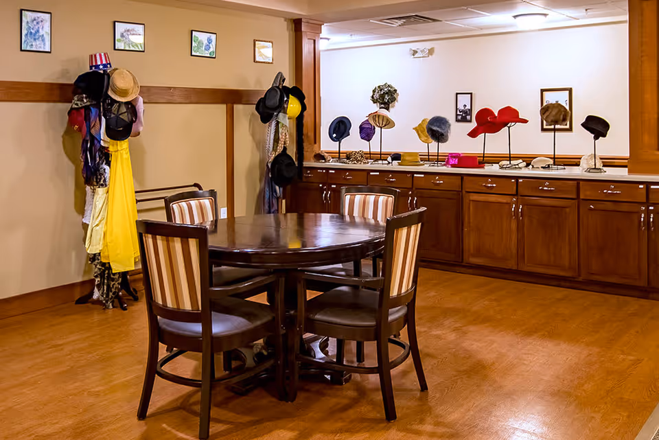 A room with a round wooden table surrounded by four chairs with striped cushions. Along the back wall, there is a long wooden cabinet displaying various hats on stands. Additional hats and scarves are hanging on coat racks to the left. The walls are decorated with small framed pictures.