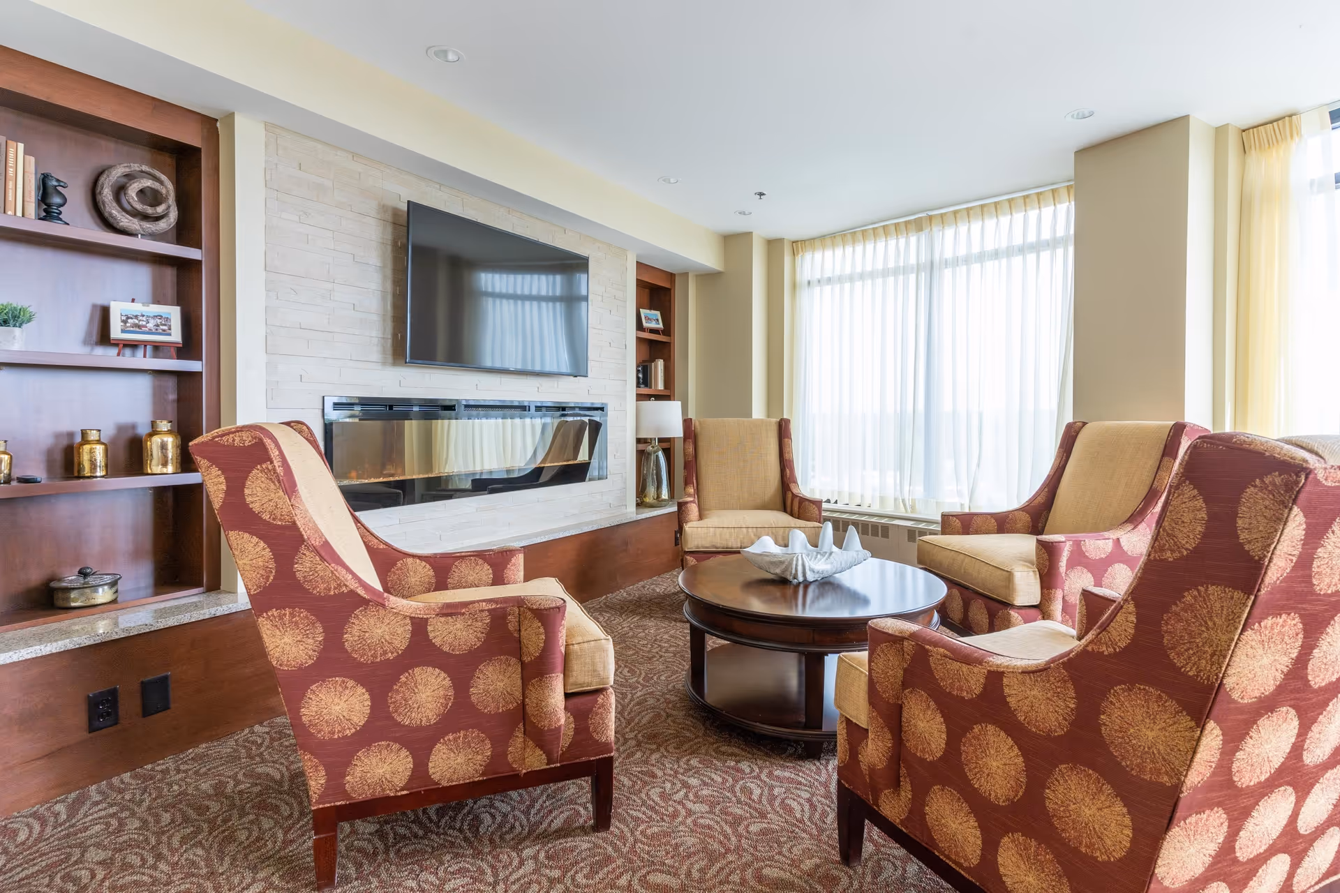 A cozy living room area with four patterned armchairs arranged around a round wooden coffee table. The room features a wall-mounted flat screen TV above a modern fireplace, built-in wooden shelves with decorative items, and large windows with sheer curtains allowing natural light to fill the space.