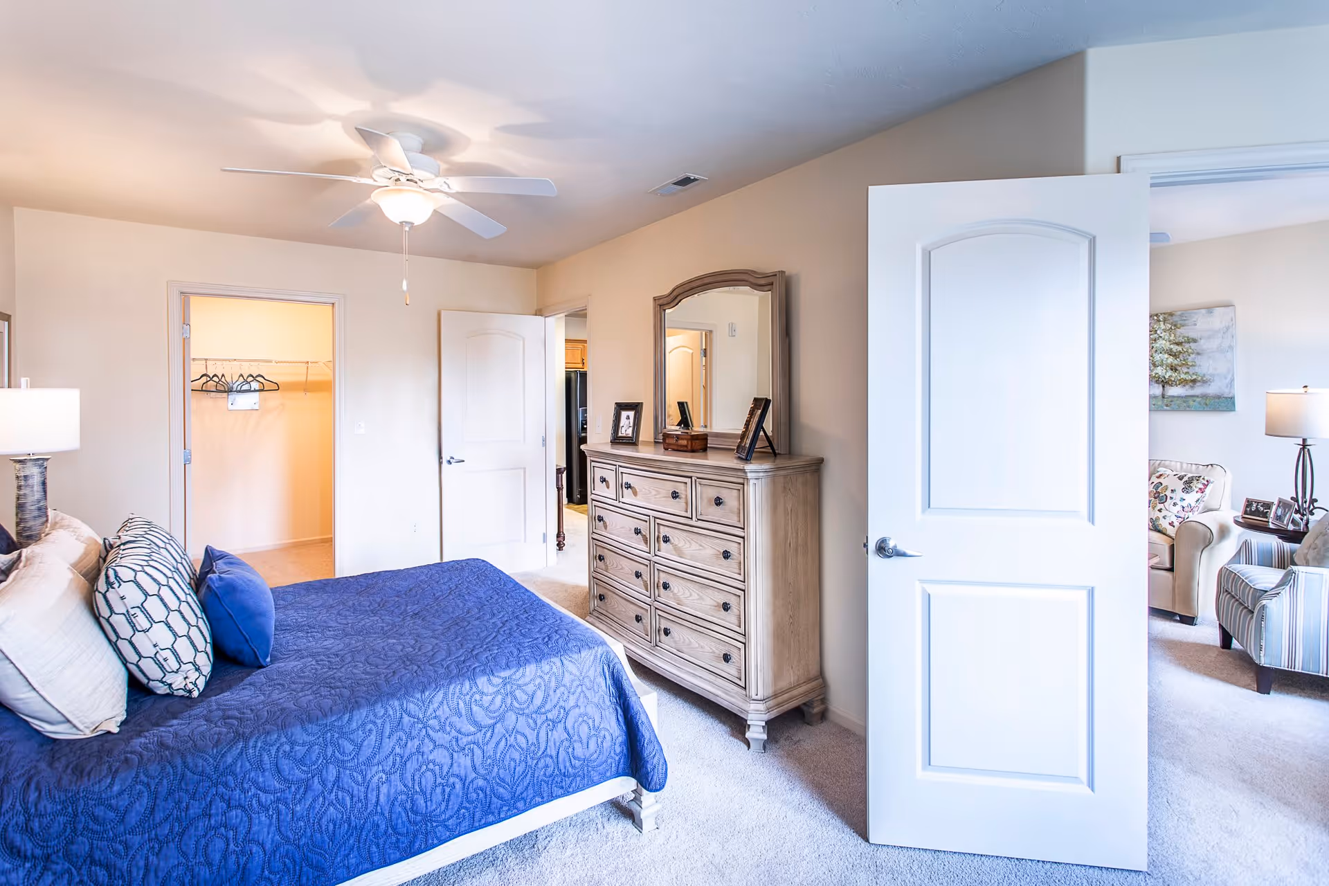 A bright bedroom with a blue bedspread on a bed, multiple pillows, a wooden dresser with a mirror, and an open door leading to a living room area with chairs and a lamp. Another door leads to a walk-in closet with hangers.