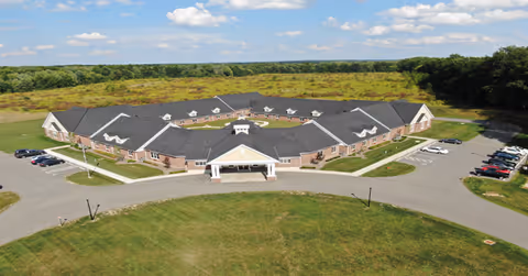 Aerial view of a single-story assisted living facility building with a large covered entrance, surrounded by parking lots and green fields under a partly cloudy sky.