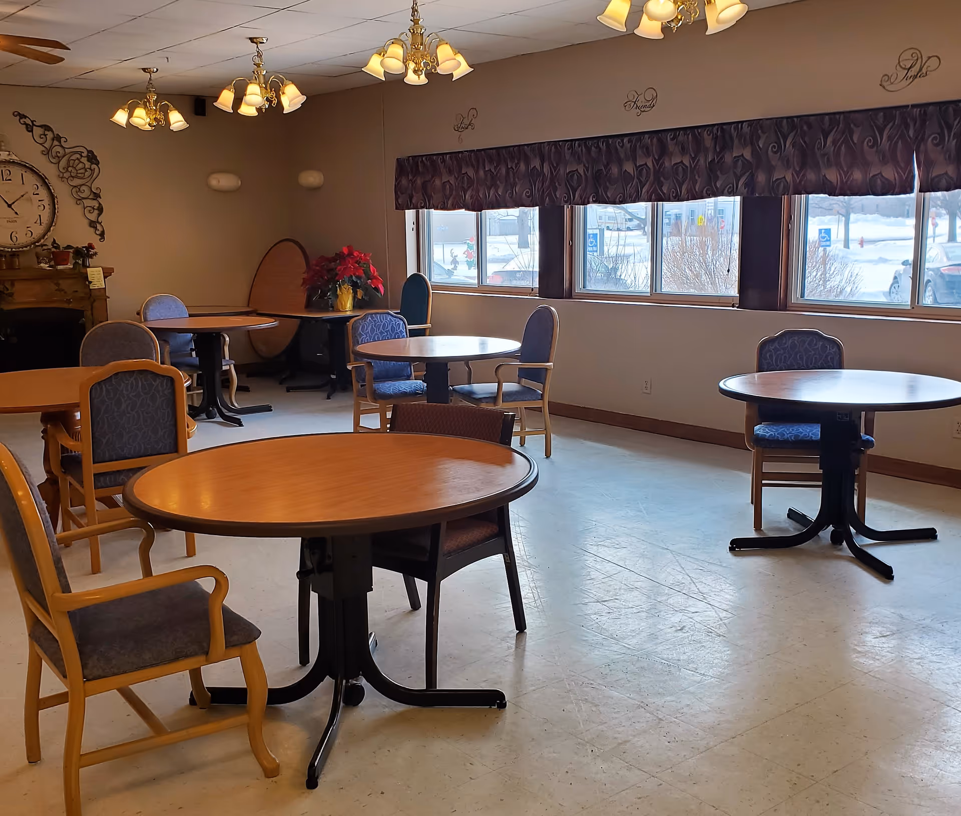 Bright communal dining room with multiple round wooden tables and upholstered chairs arranged near windows.