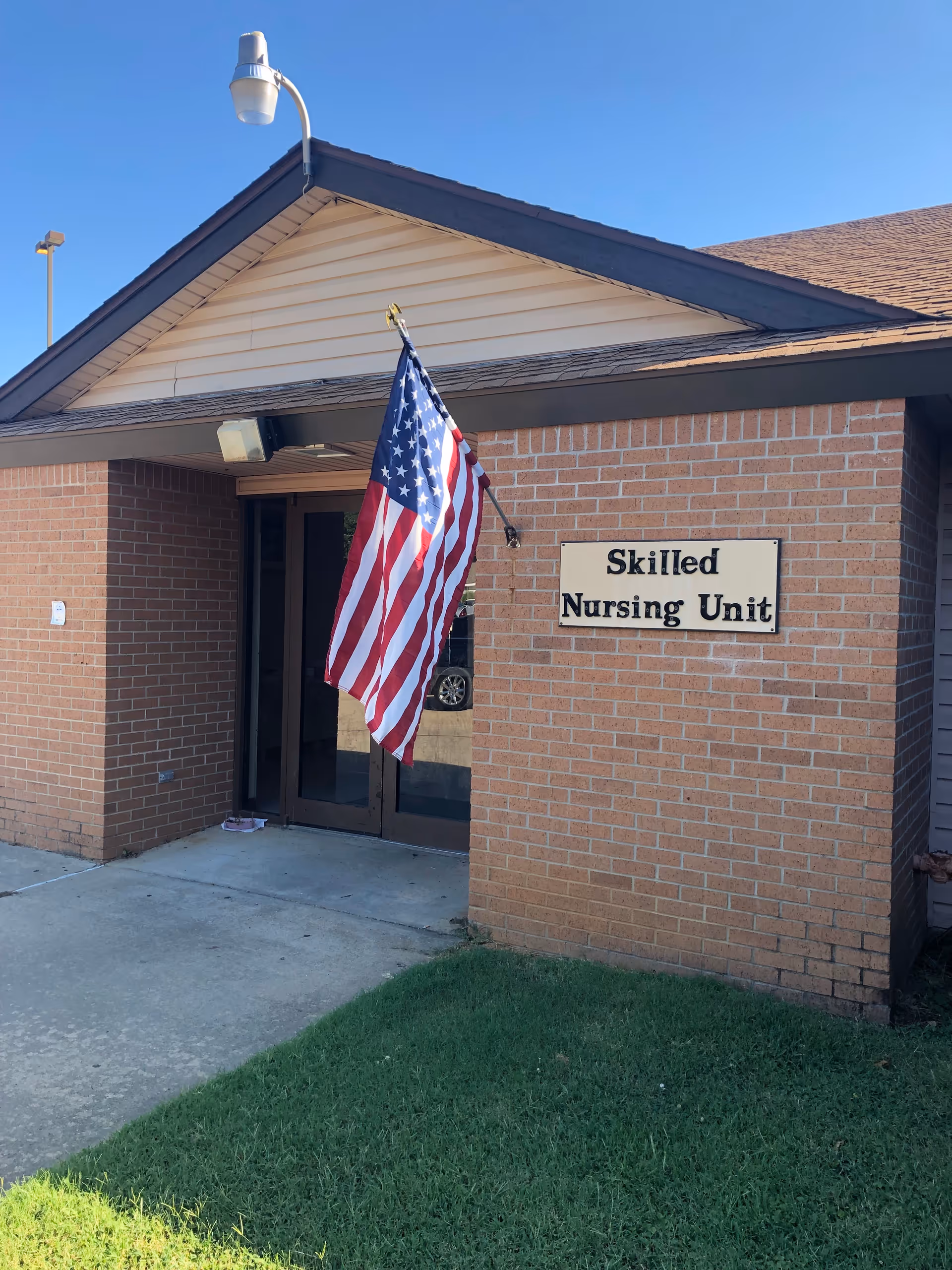 Entrance to a skilled nursing unit with a brick exterior wall, a sign that reads 'Skilled Nursing Unit', and an American flag mounted on the wall near the door. The door is glass with a dark frame, and there is a small patch of grass in front of the building.