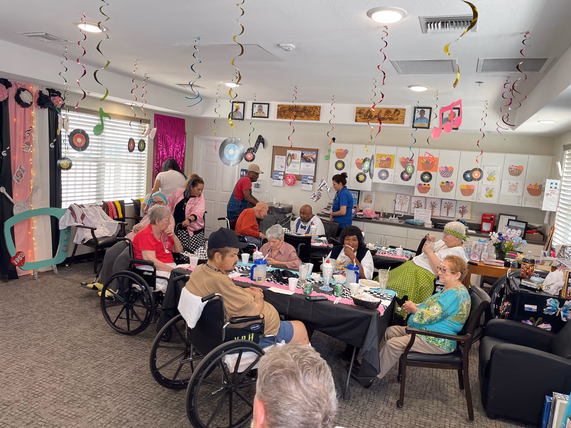 Residents seated around decorated tables in a senior living common/dining room with hanging streamers and craft decorations.