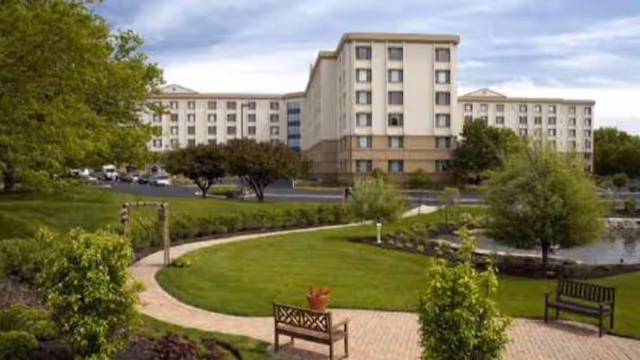 Landscaped courtyard with benches, a pond and a paved walkway in front of a multi-story retirement community building.