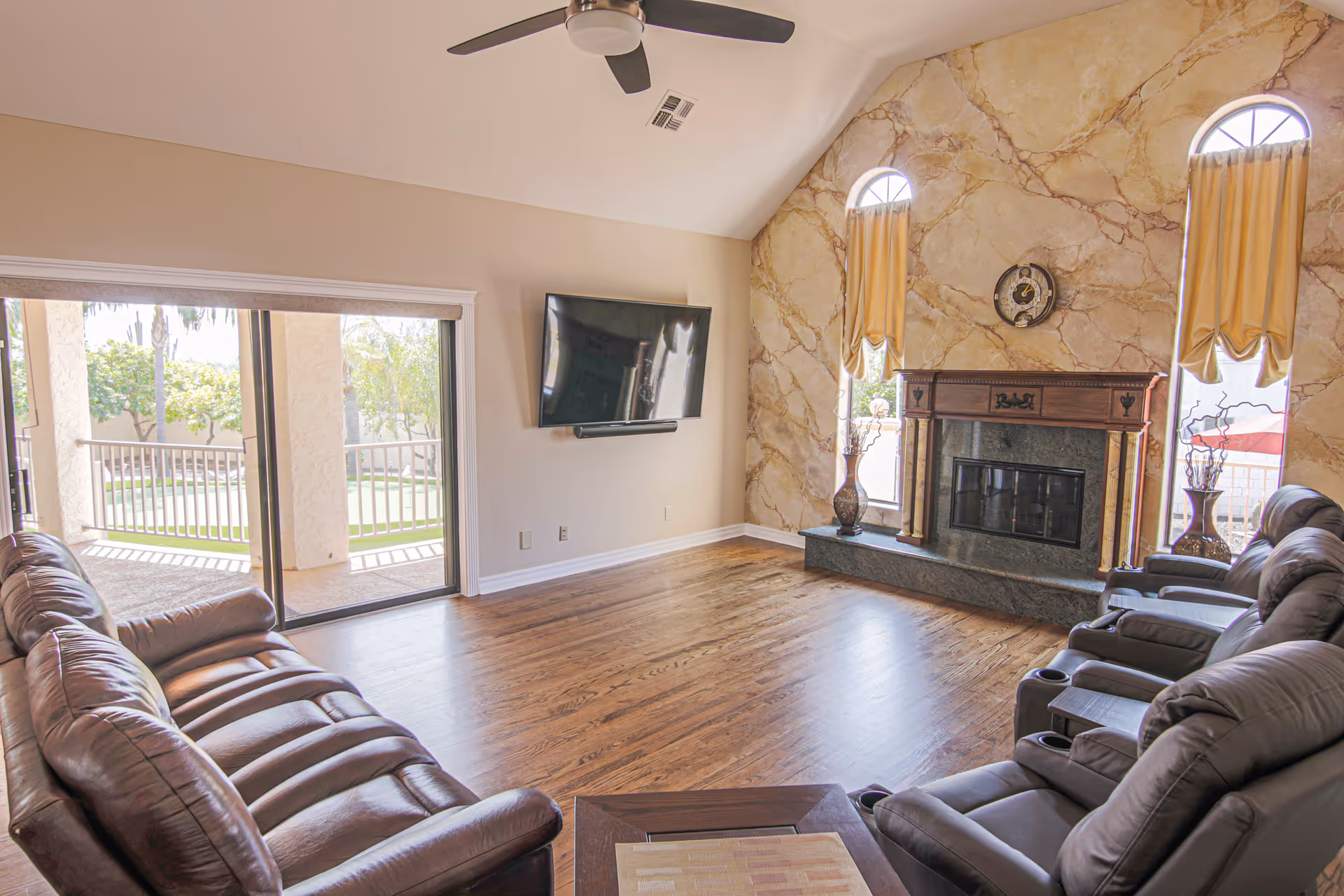Sunlit living room with leather recliners, a wall-mounted TV, a decorative fireplace, and sliding glass doors to a patio.