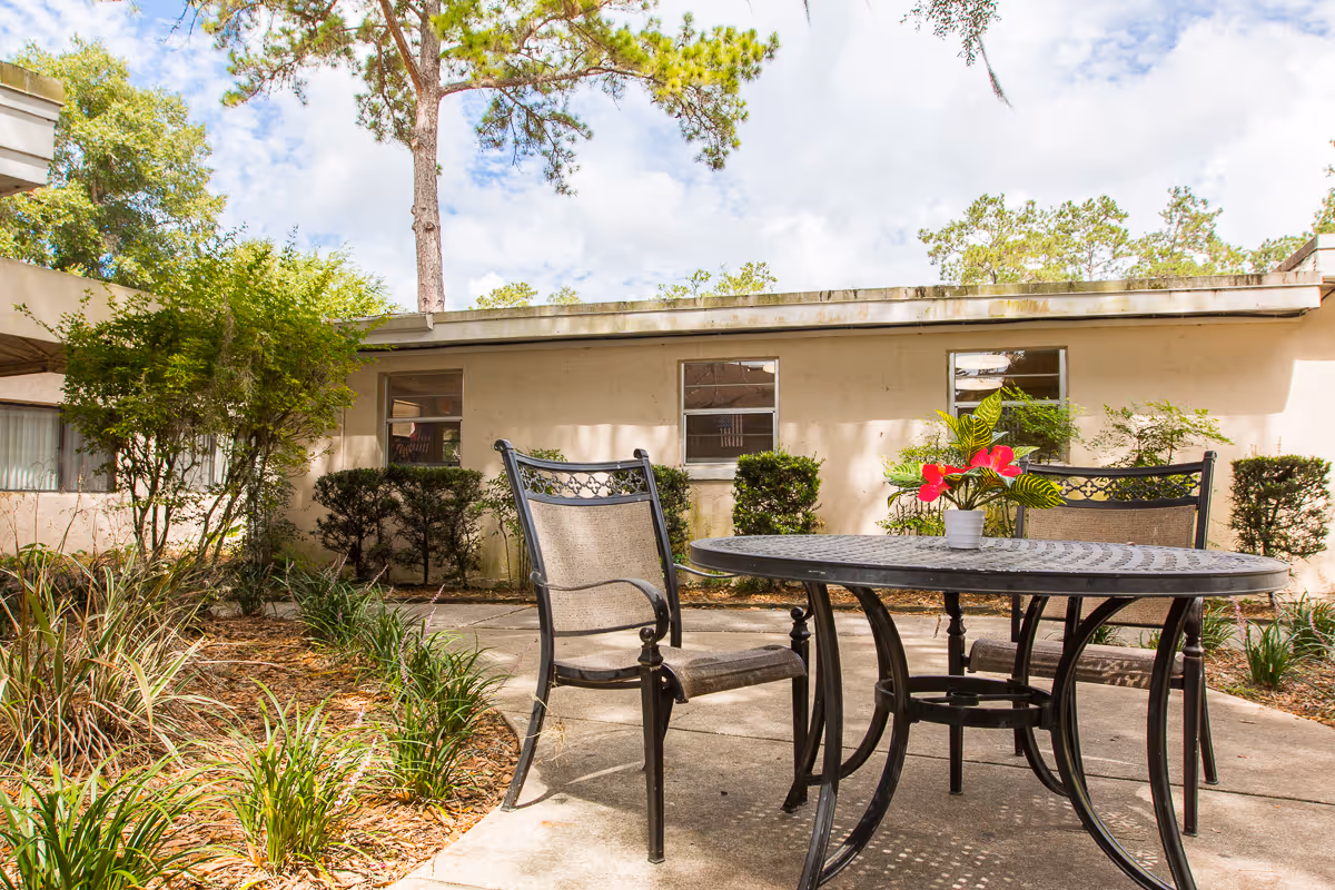 Outdoor patio area at Signature HealthCARE of Gainesville with a round metal table and two chairs. A small potted plant with red flowers is placed on the table. The patio is surrounded by greenery, bushes, and trees, with a beige building wall and windows in the background under a partly cloudy sky.
