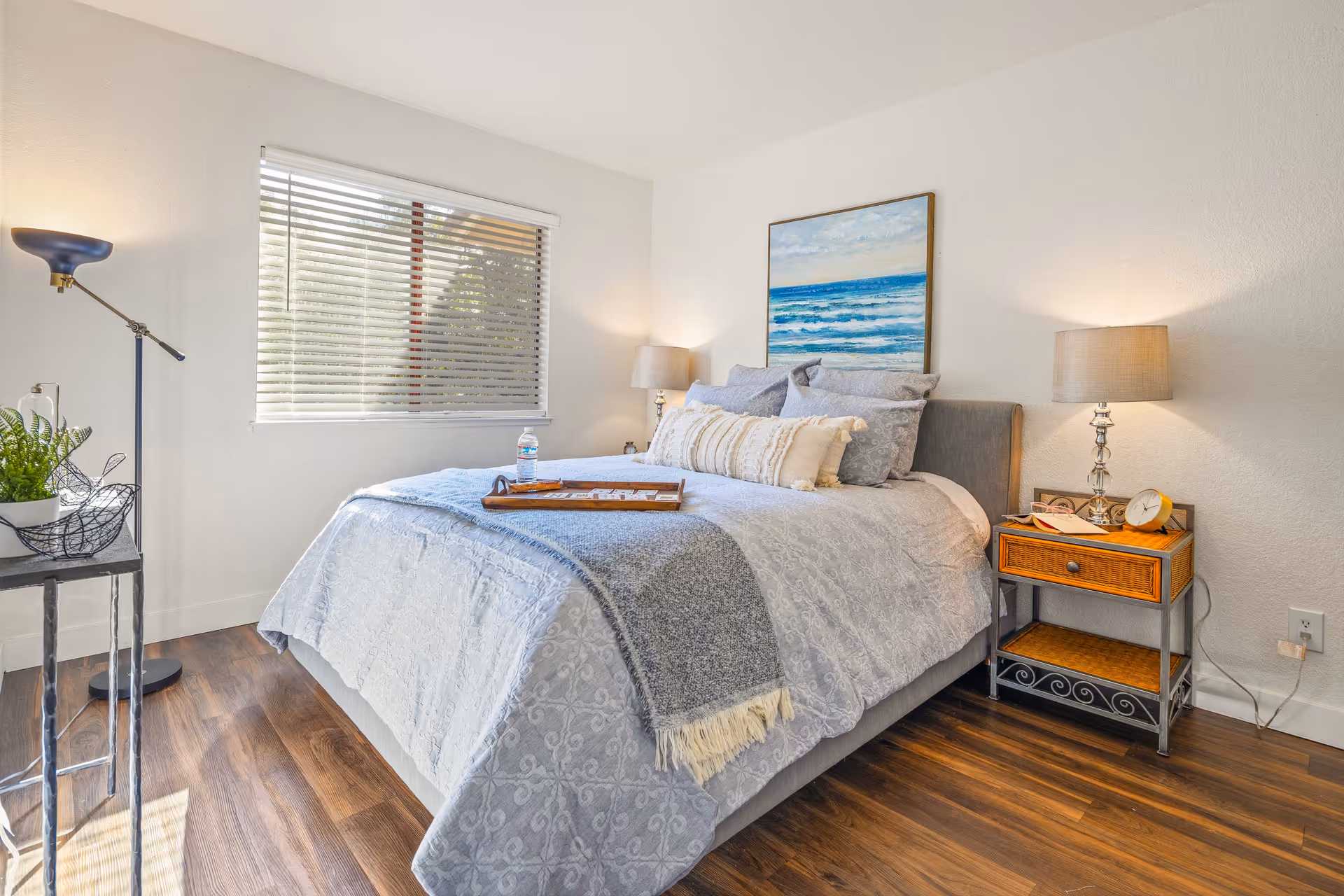 A bright bedroom with a neatly made bed featuring light blue and gray bedding, a wooden nightstand with a lamp, clock, and picture frame, a floor lamp, a small table with a plant, and a window with blinds letting in natural light. A painting of an ocean scene hangs above the bed.