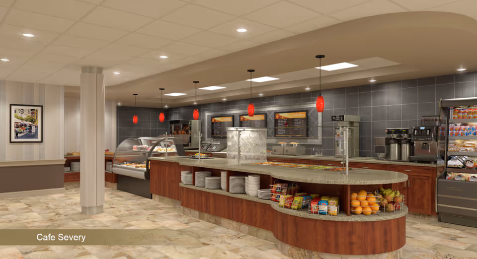 Interior view of a cafe area with a curved serving counter displaying plates, snacks, and fruits. Behind the counter are food warmers, coffee machines, and menu boards on a tiled wall. The floor is tiled, and there are red pendant lights hanging from the ceiling.