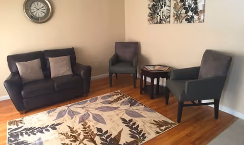 A cozy living room area with a dark brown loveseat with two light-colored cushions, two gray armchairs, a small wooden side table with magazines, a wall clock, and two framed botanical prints on the wall. The floor is hardwood with a decorative area rug featuring a leaf pattern in shades of brown and purple.