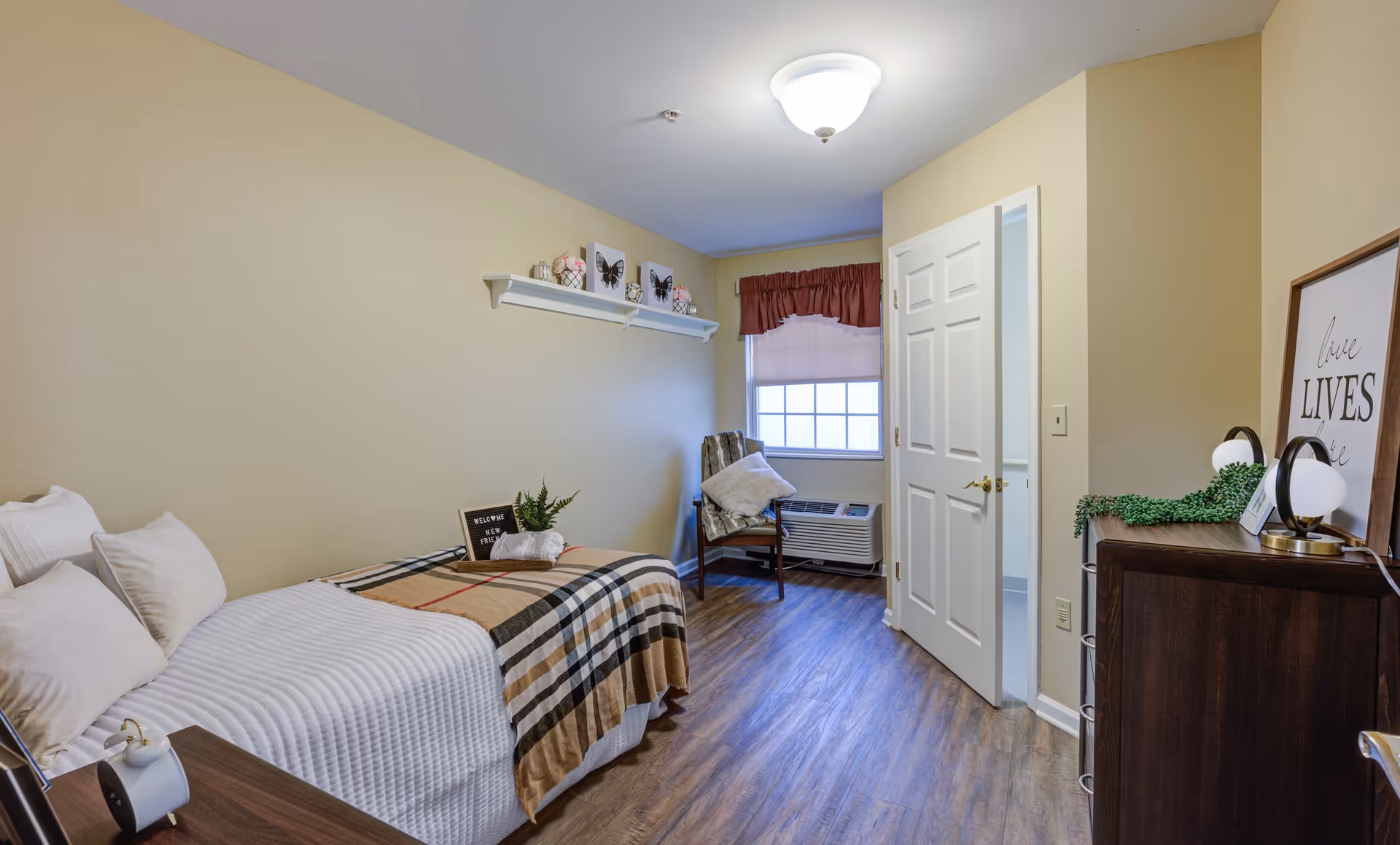 A small, cozy bedroom with a single bed covered in white and plaid bedding. There are three pillows on the bed and a small plant on the bed. A wooden chair with a cushion is placed near a window with a red valance and white blinds. A white door is open, revealing a glimpse of an adjacent room. A dark wooden dresser with a lamp and framed artwork is on the right side of the room. The floor is wood, and the walls are painted a light beige color.