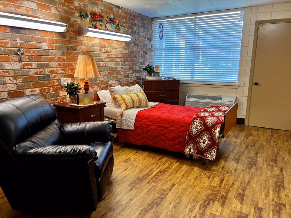 A cozy bedroom with a single bed topped with a red quilt, a leather recliner, nightstand with a lamp, exposed brick wall and window blinds.