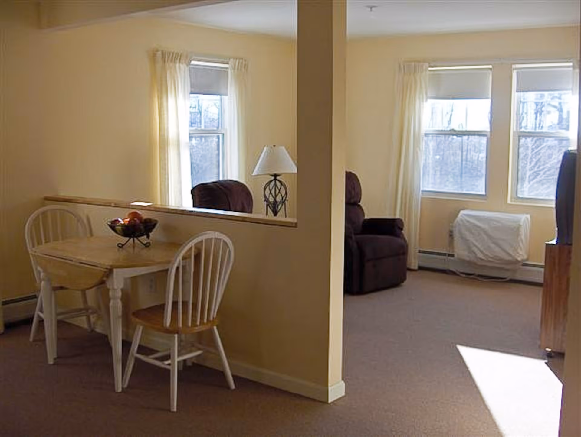Interior view of a living space divided by a half wall. On the left side, there is a small wooden dining table with two chairs and a bowl of fruit. On the right side, there is a brown recliner chair, a lamp on a small table, two windows with white curtains, and a covered radiator beneath the windows.