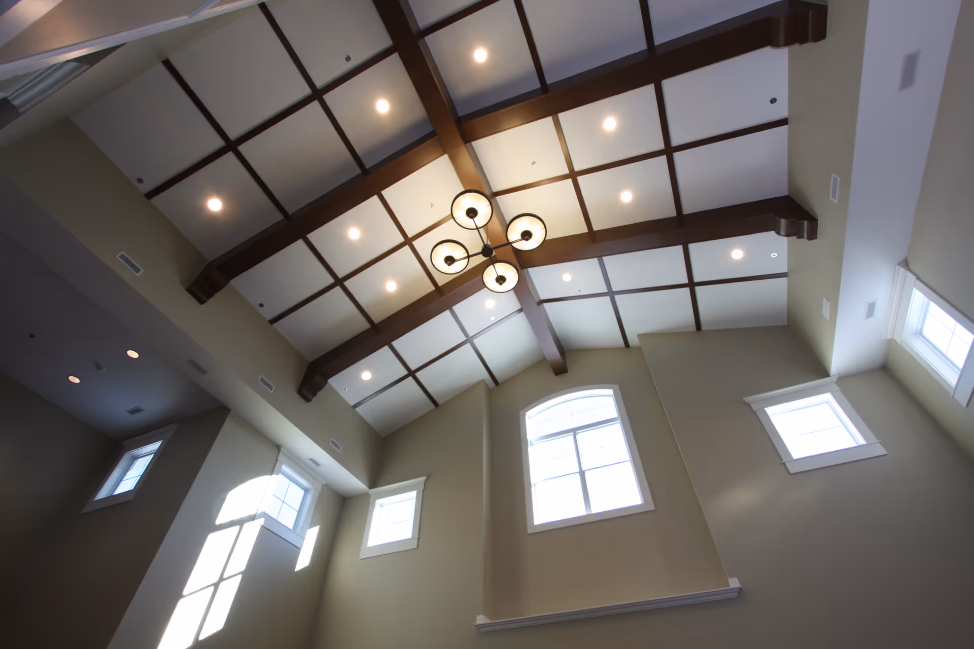 Vaulted interior ceiling with coffered wood beams, a central chandelier, recessed lighting, and tall arched windows.