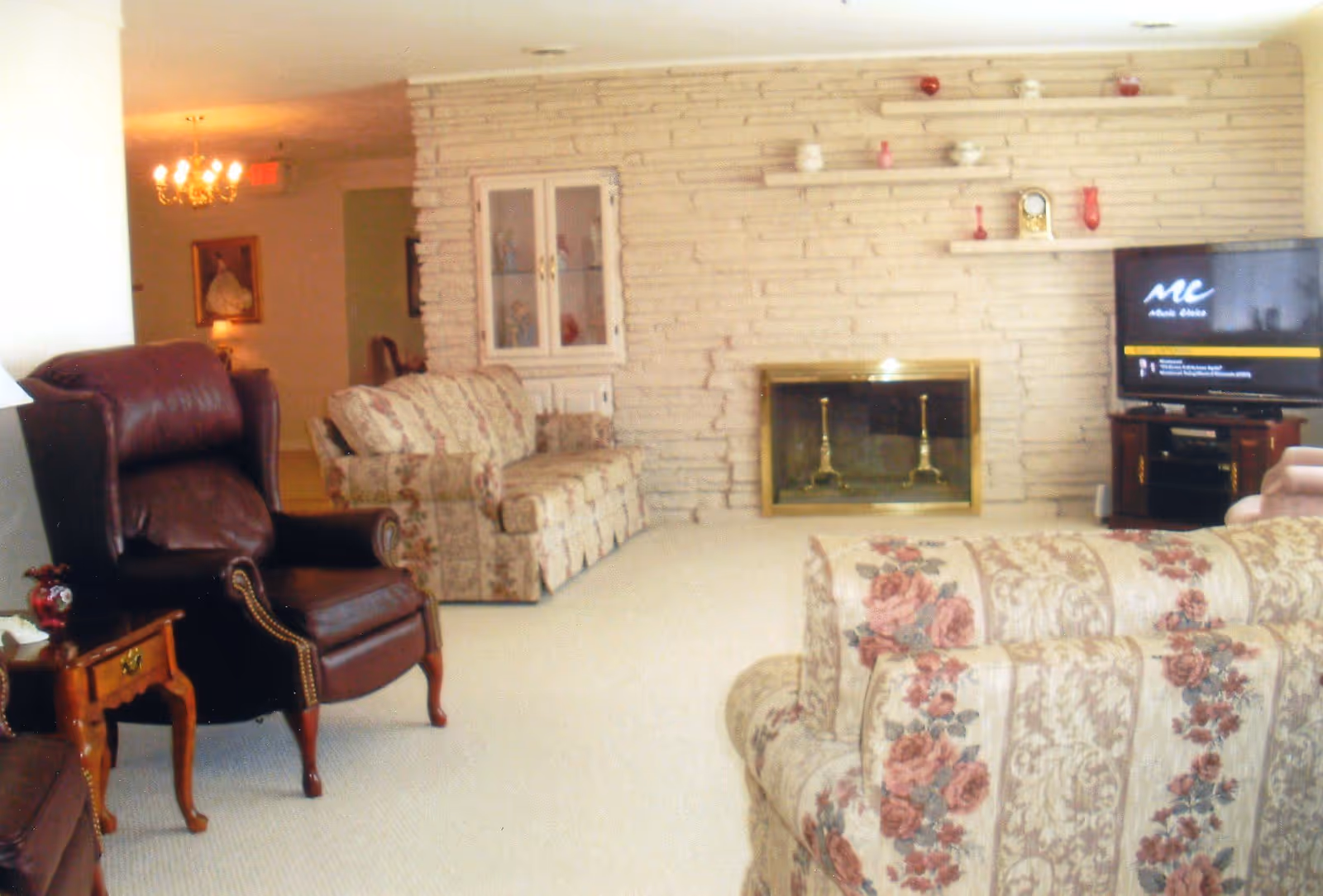 A living room with floral patterned sofas and a dark brown leather armchair. The room features a stone wall with a fireplace and shelves holding decorative items. A television is placed on a wooden stand to the right of the fireplace. There is a chandelier and a framed picture visible in the background.