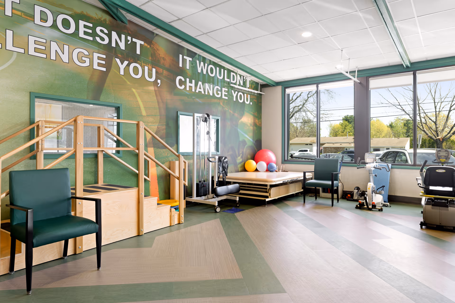 A bright rehabilitation room with large windows letting in natural light. The room features exercise equipment including a stationary bike, a set of wooden stairs with handrails for physical therapy, a padded platform with colorful exercise balls, and two green chairs. The wall has a motivational quote partially visible that reads, 'IT DOESN'T CHALLENGE YOU, IT WOULDN'T CHANGE YOU.'