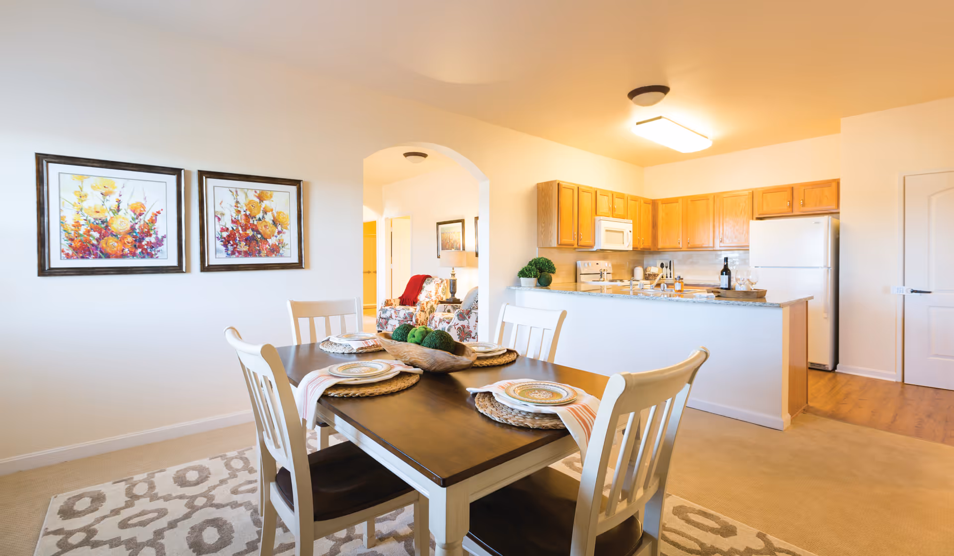 Bright and cozy dining area with a wooden table set for four, featuring plates and napkins. The dining area is adjacent to a kitchen with wooden cabinets, a refrigerator, microwave, and stove. Two colorful floral paintings hang on the wall, and an archway leads to a living room with floral armchairs and a lamp.