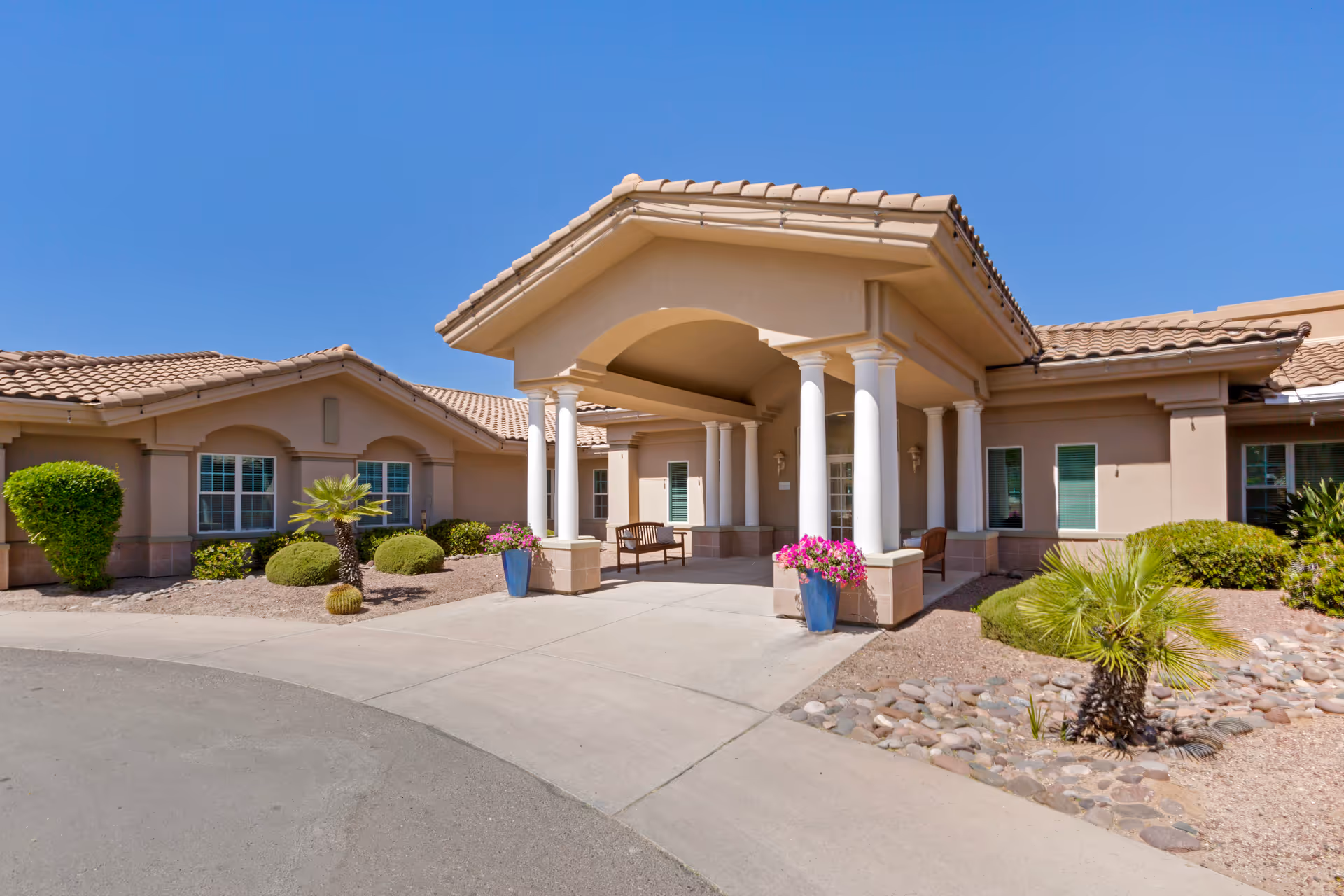 Front entrance of a single-story stucco building with a covered porte-cochère supported by white columns, benches, potted flowers and desert landscaping.