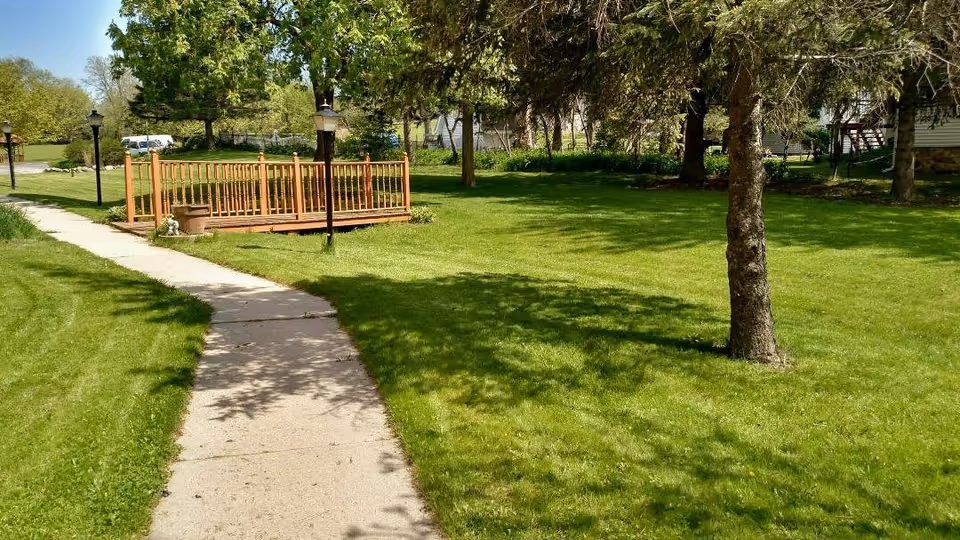 A sunny outdoor area with a concrete pathway leading to a small wooden bridge surrounded by green grass and trees. There are several lamp posts along the pathway and a house partially visible on the right side.