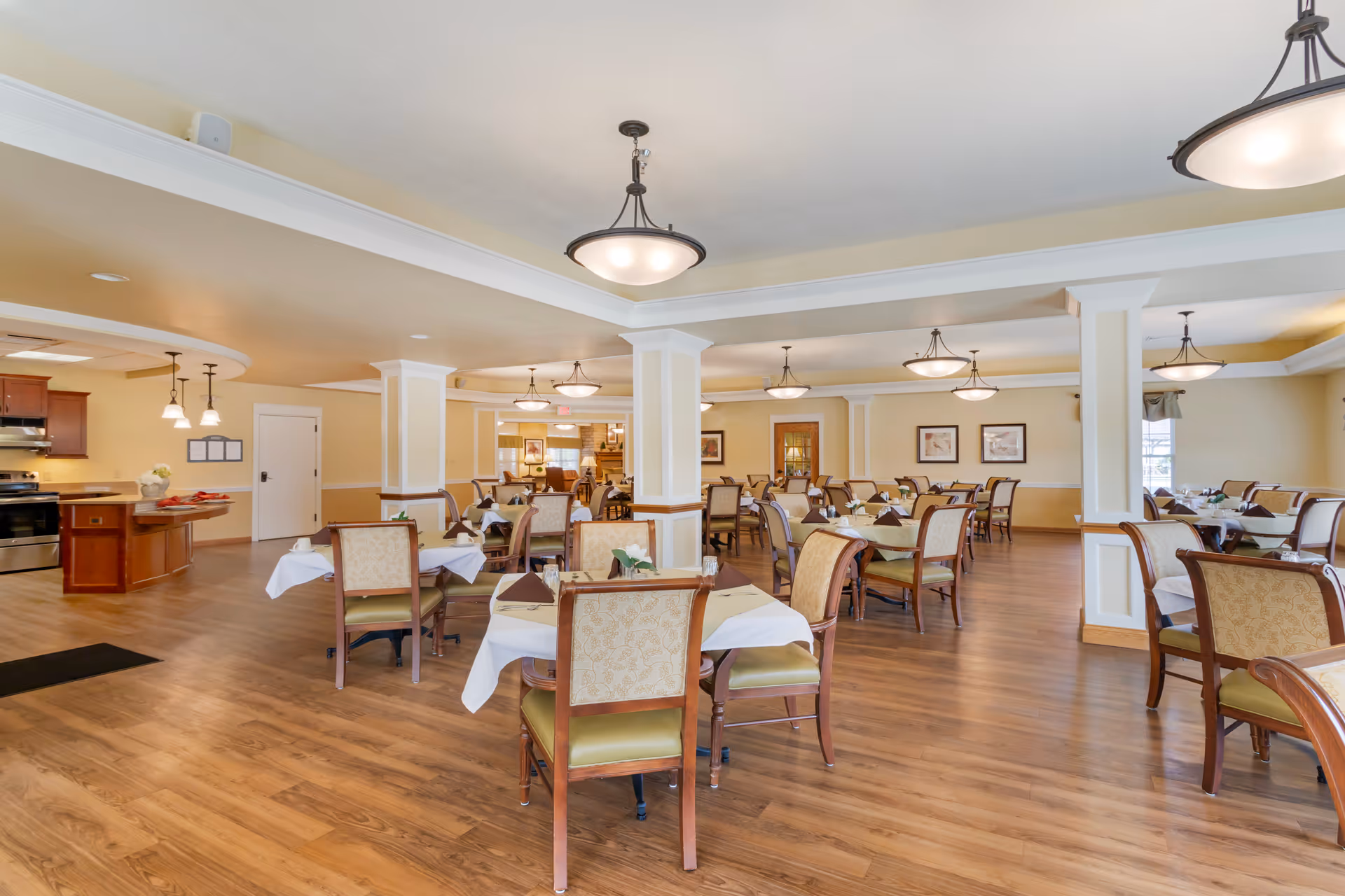 A spacious dining room with multiple tables covered with white tablecloths and set with napkins, glasses, and small flower arrangements. The room has wooden flooring, beige walls with white trim, and several ceiling light fixtures. In the background, there is a kitchen area with wooden cabinets and stainless steel appliances.