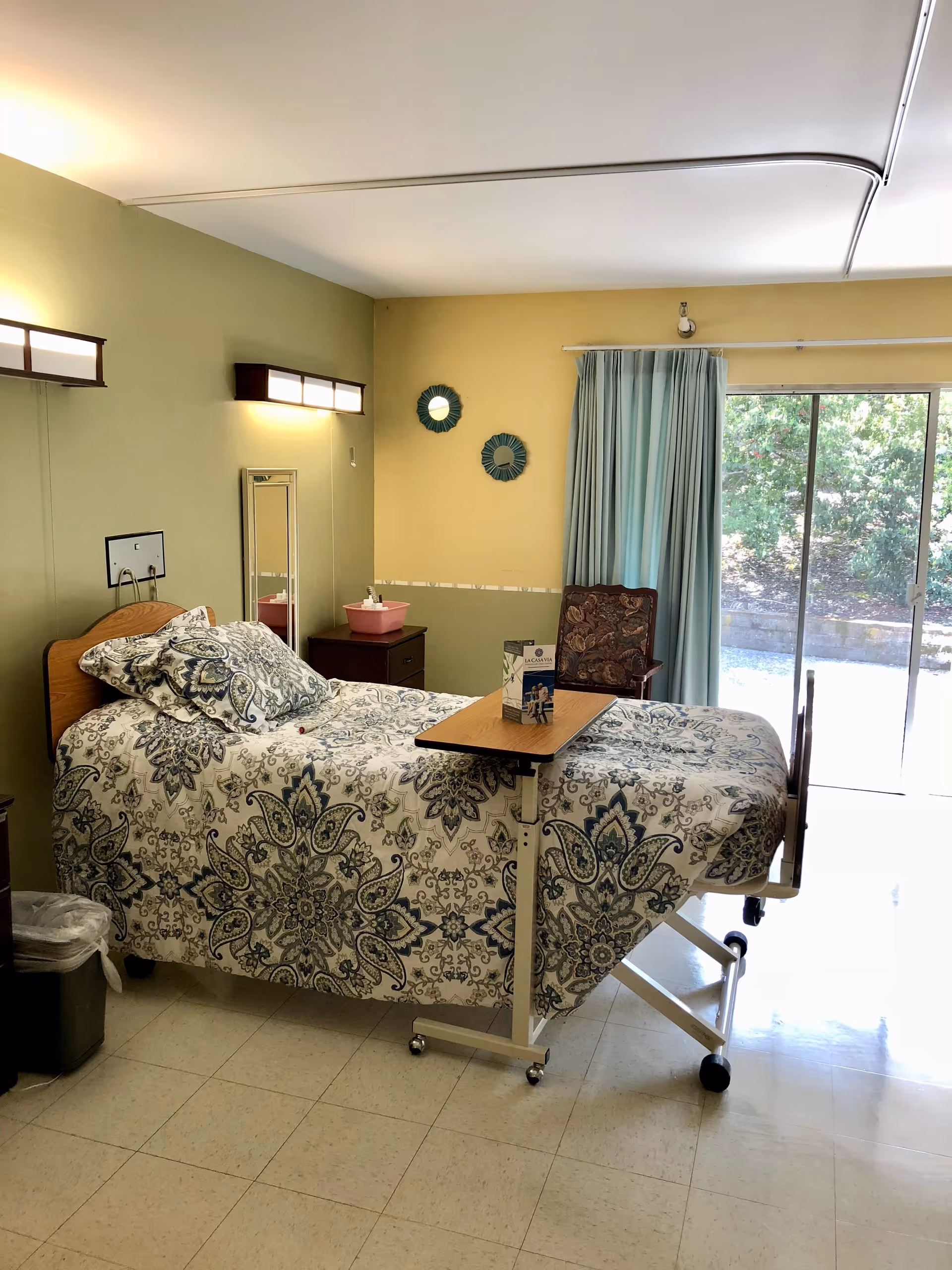 A single hospital-style bed with a patterned bedspread in a room with light green walls. The bed has an overbed table with a brochure on it. There is a wooden chair with a floral cushion near a window with light blue curtains. The window is a sliding glass door that opens to an outdoor area with greenery. The room has two wall-mounted lights and a small mirror above a nightstand with a pink container on it.