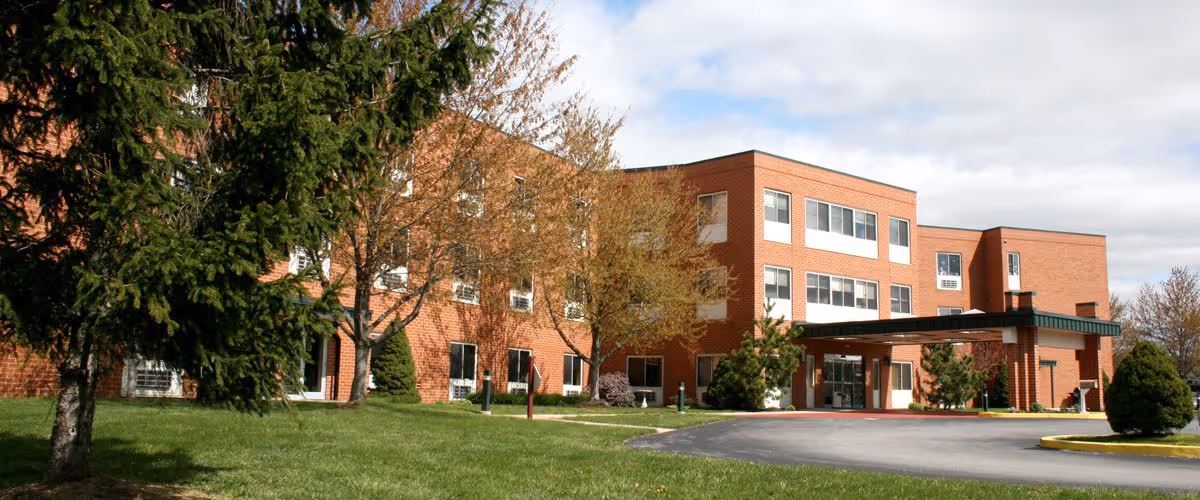 Exterior view of a three-story brick building with multiple windows, surrounded by trees and a grassy lawn under a partly cloudy sky. The building has a covered entrance with a driveway in front.