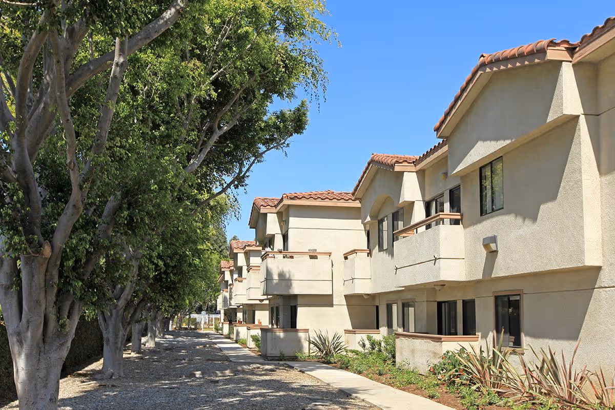 Row of stucco, tile-roofed residential buildings beside a tree-lined walkway under a clear blue sky.