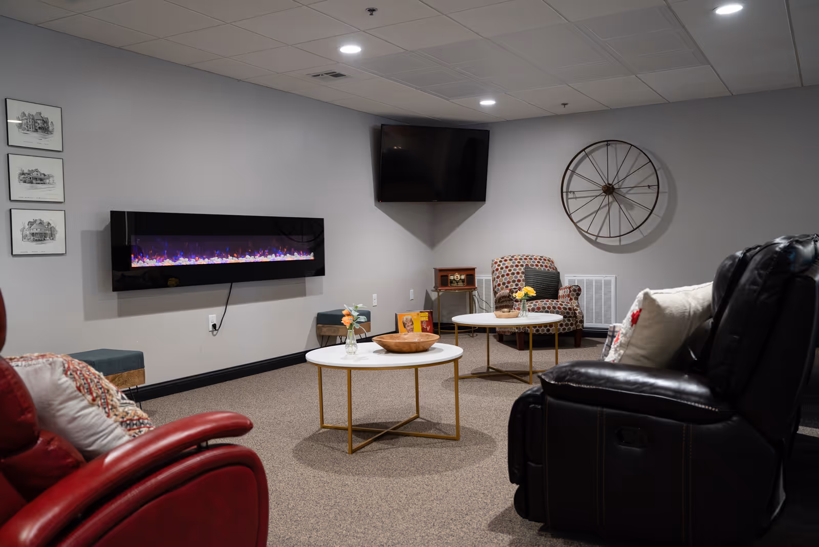 A cozy living room area with a modern electric fireplace mounted on a gray wall, a flat-screen TV in the corner, two round white coffee tables with gold legs, a patterned armchair, and two recliner chairs in red and black leather. The room has a carpeted floor, recessed ceiling lights, and decorative wall art including three framed sketches and a large metal wheel.