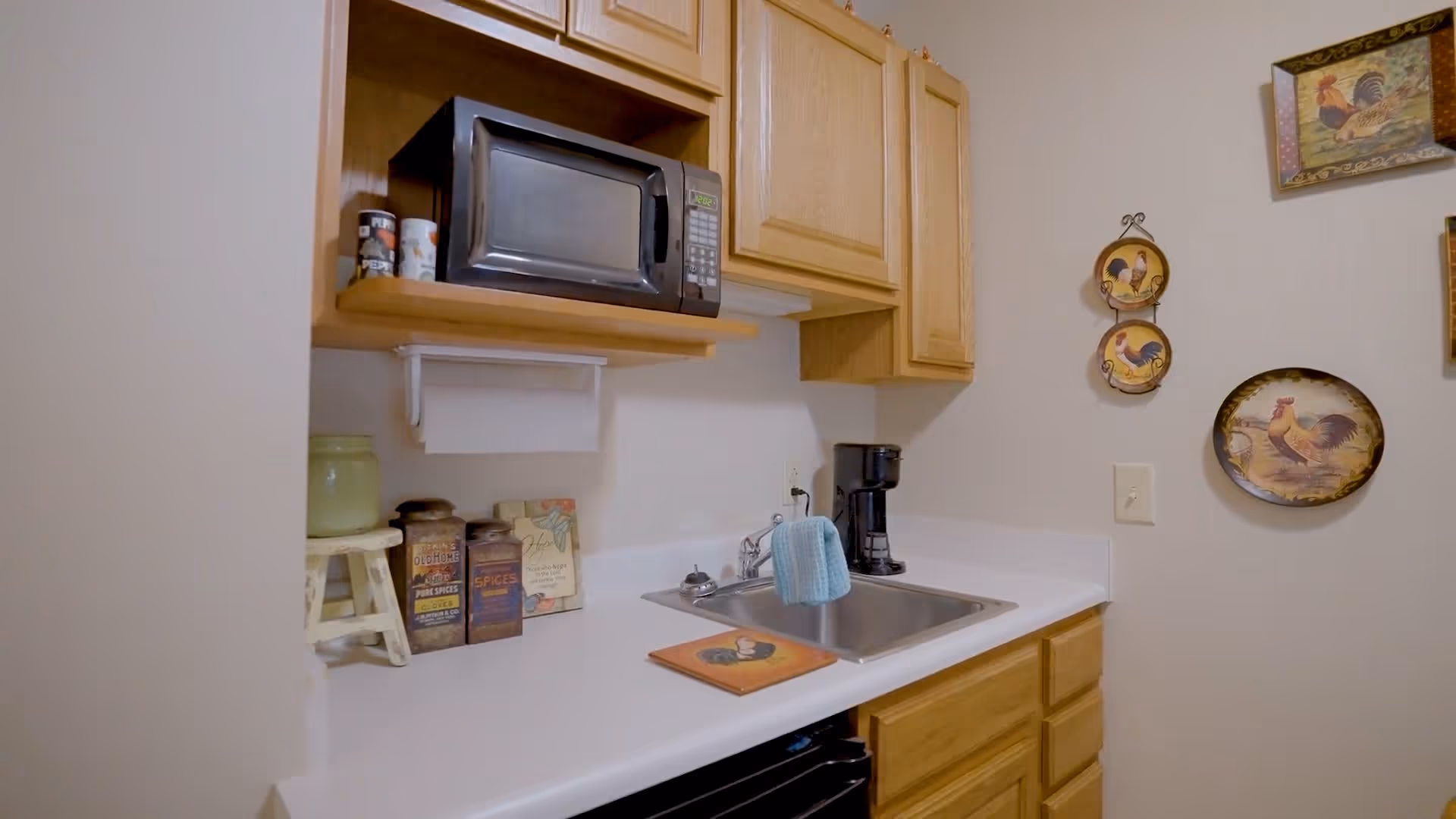 A small kitchen area with wooden cabinets, a microwave on a shelf, a coffee maker next to a sink with a blue towel hanging on the faucet, and decorative rooster-themed plates and artwork on the wall.