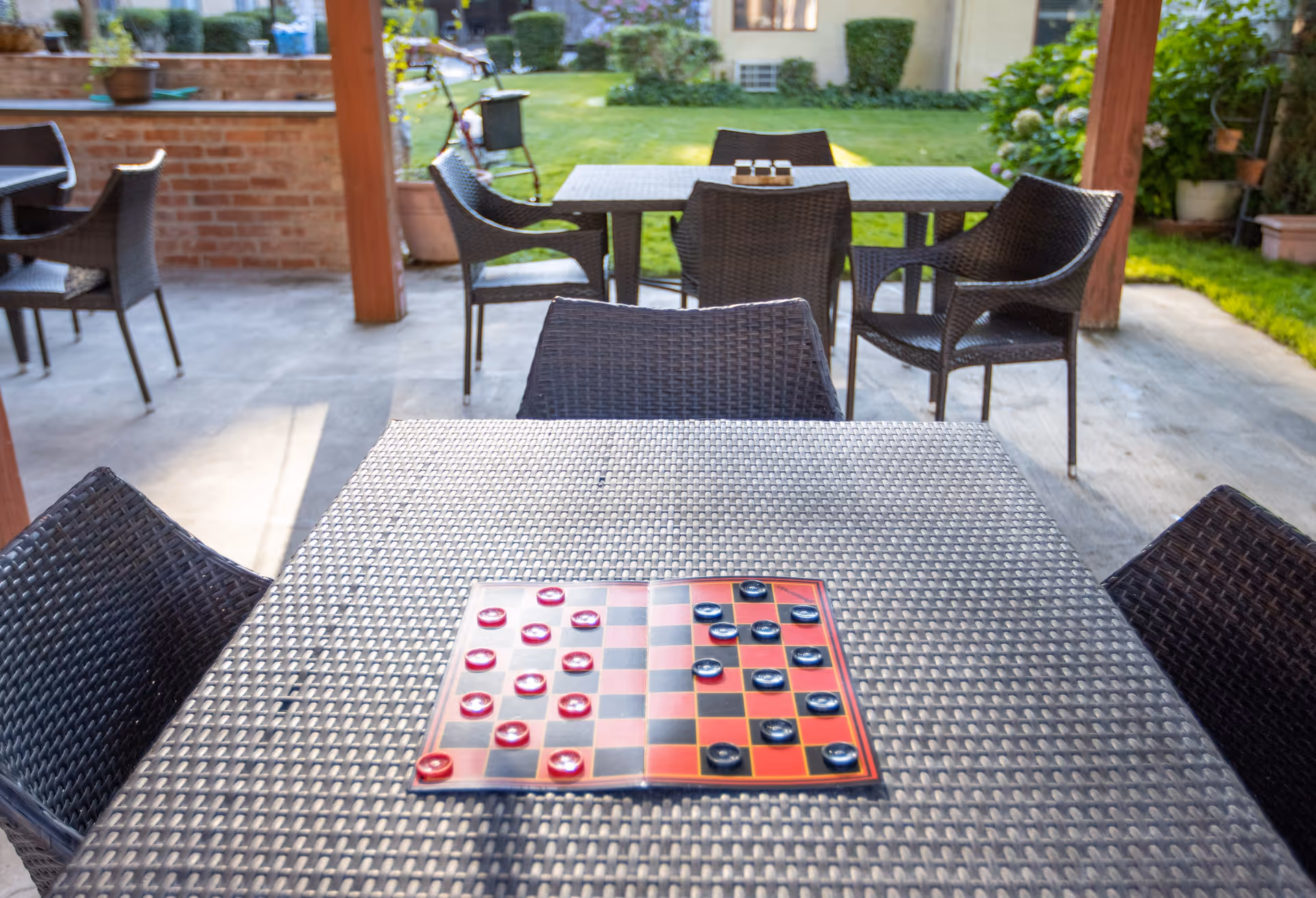 Outdoor patio area with multiple wicker chairs and tables. A checkers board with red and black pieces is set up on the closest table. The patio is covered and overlooks a grassy garden area with plants and a building in the background.