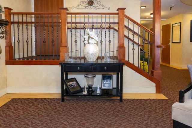 Lobby or common area showing a wooden staircase railing behind a black console table topped with a decorative vase of flowers.