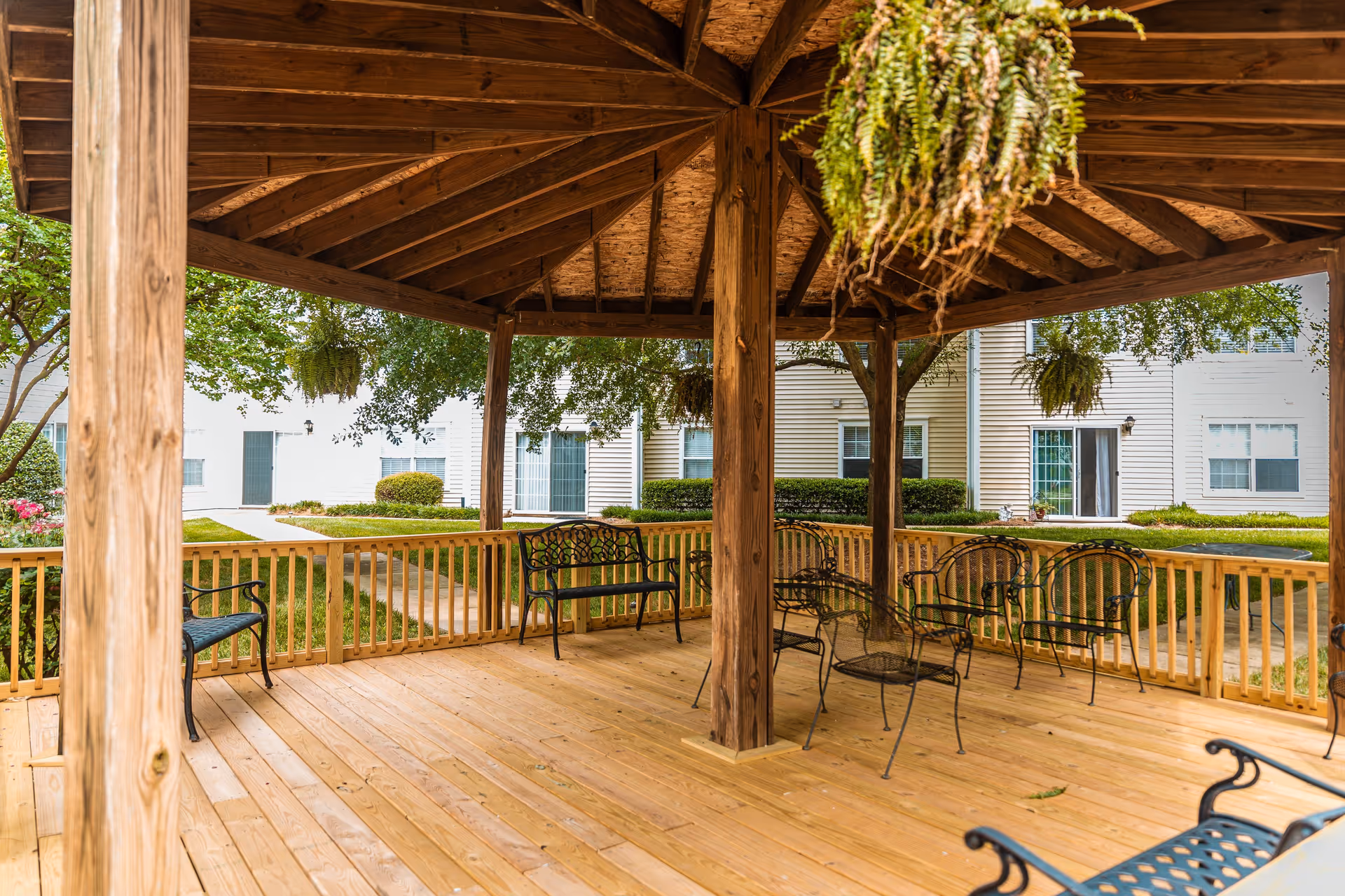 Wooden gazebo with metal chairs and hanging ferns overlooking a grassy courtyard and adjacent apartment building.