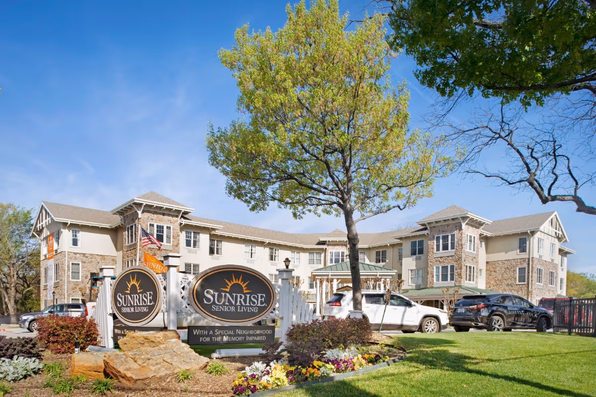 Exterior view of Sunrise Senior Living facility with a large sign in front that reads 'Sunrise Senior Living' and 'With a Special Neighborhood for the Memory Impaired'. The building is three stories tall with stone and beige siding, surrounded by green grass, trees, and parked cars under a clear blue sky.