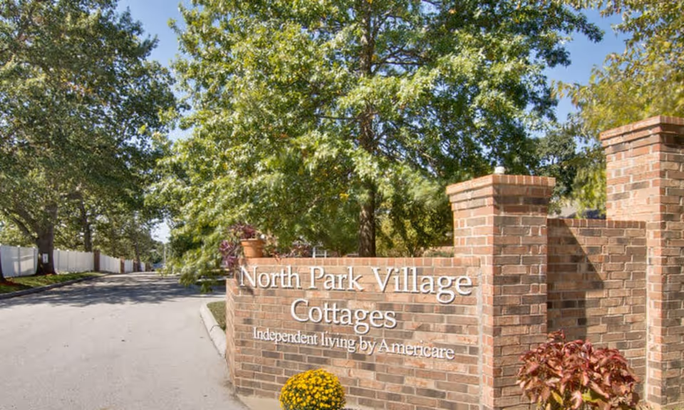 Brick entrance sign reading 'North Park Village Cottages' beside a tree-lined driveway.