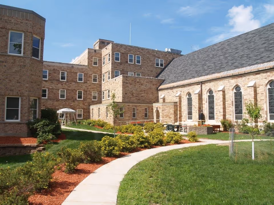 Landscaped courtyard with a curved sidewalk, lawn, and outdoor seating beside a multi-story brick assisted living building.
