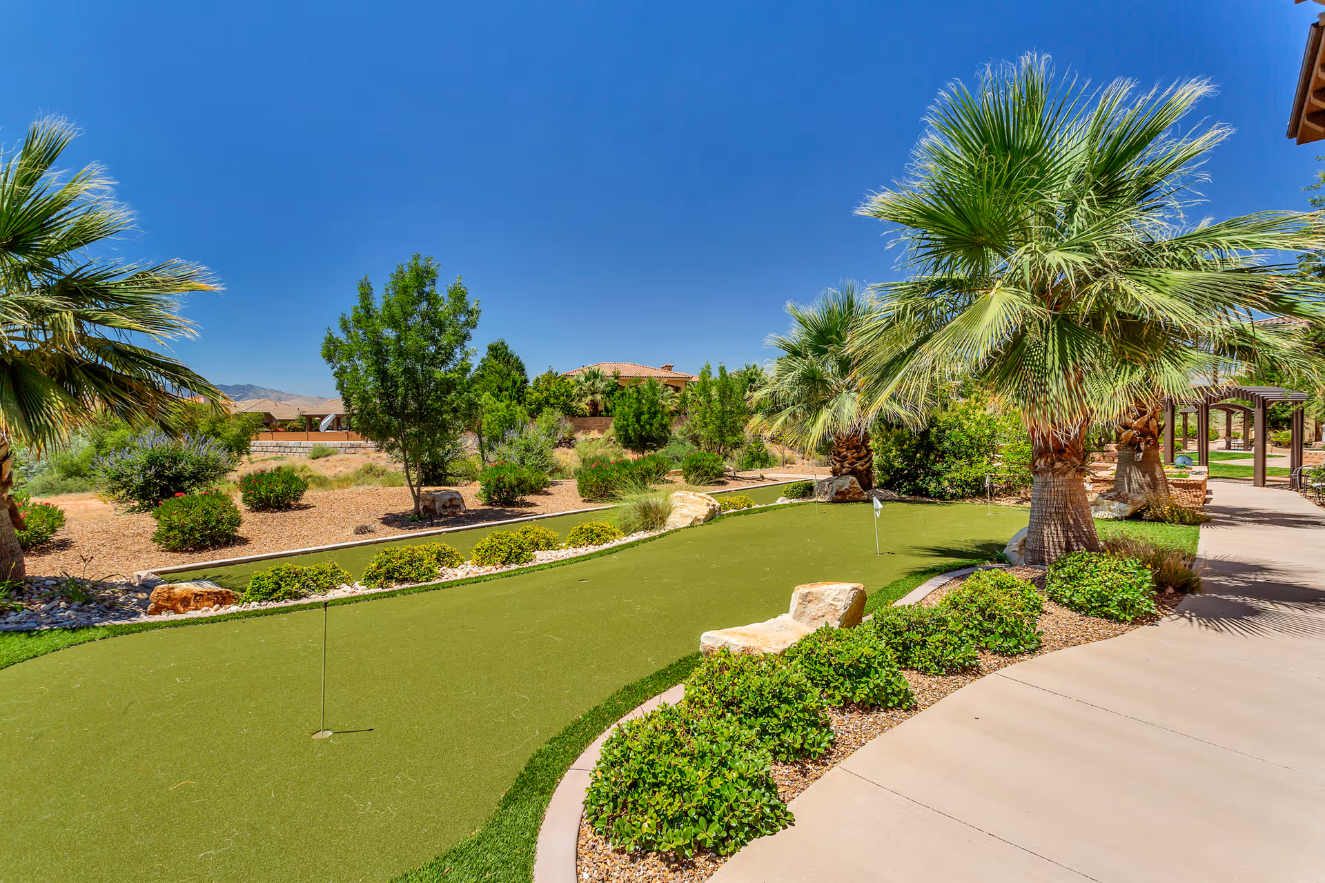 Outdoor putting green surrounded by palm trees, shrubs, and a paved walkway under a clear blue sky at The Retreat at Sunbrook.