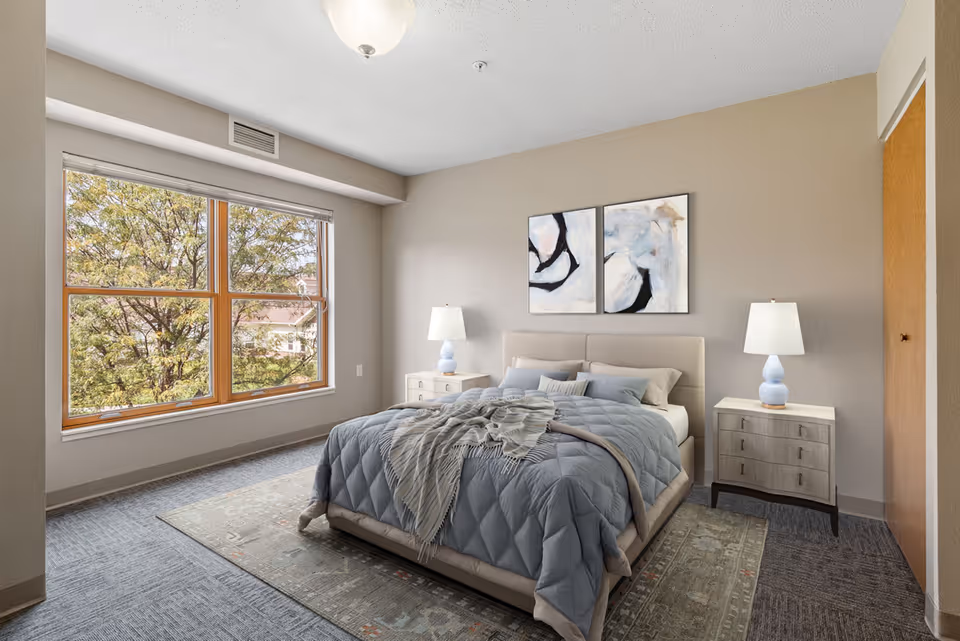 A bright bedroom with a large window showing trees outside. The room features a neatly made bed with a blue quilt and multiple pillows, two bedside tables each with a lamp, a patterned rug under the bed, and two abstract paintings on the wall above the headboard.