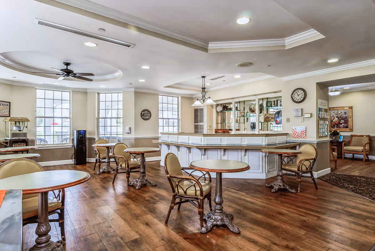 A bright and spacious common area with wooden floors, round tables with chairs, a popcorn machine, a water dispenser, and a white counter with glass shelves behind it. Large windows allow natural light to fill the room. The ceiling has recessed lighting and a ceiling fan. A clock and some wall decorations are visible.