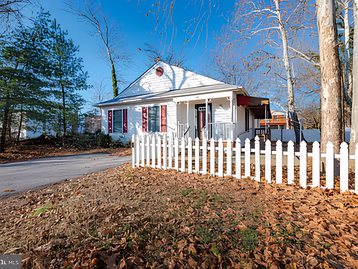 A white single-story house with red shutters and a small front porch, surrounded by a white picket fence. The yard is covered with dry leaves and there are trees with bare branches around the house under a clear blue sky.