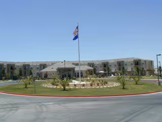 Exterior view of Portofino Senior Apartments showing a large, curved three-story building with multiple windows, a central entrance with a covered drop-off area, a flagpole with the American flag and another flag, and landscaped greenery in front under a clear blue sky.