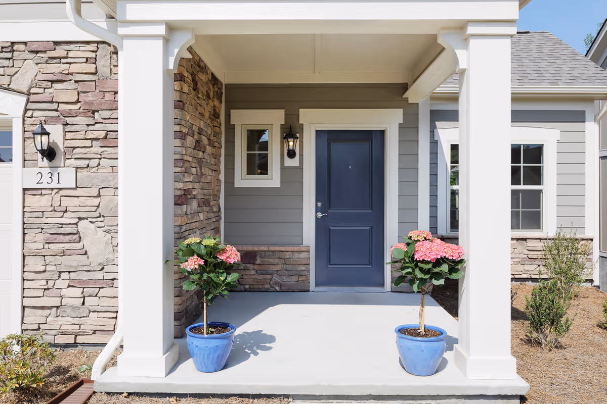 Front porch of a residential unit with a dark blue door, two white pillars, stone and gray siding walls, two blue pots with pink flowering plants, and a house number 231 on the left side.