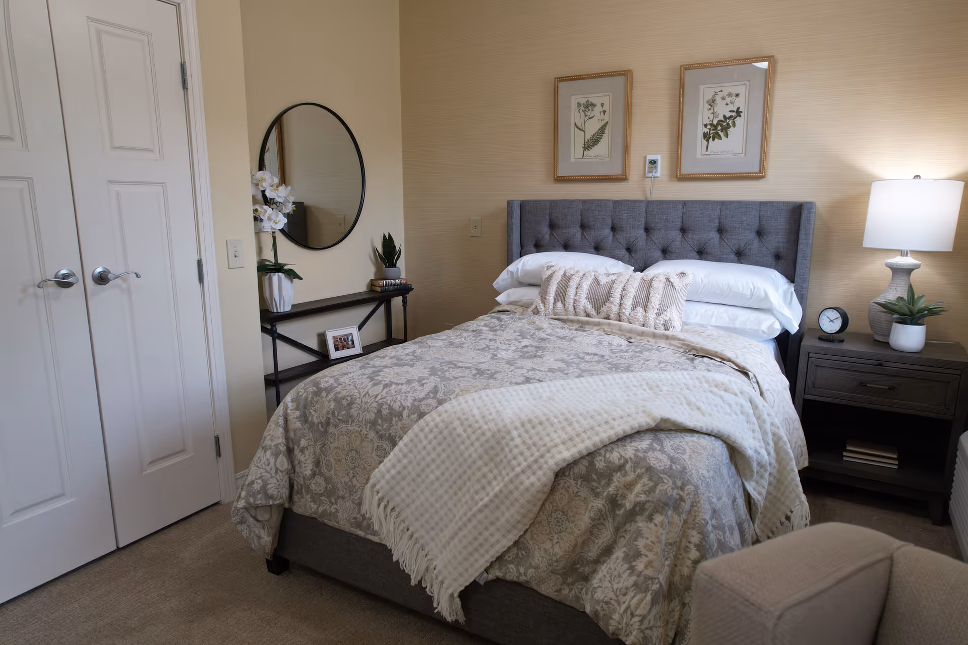 Well-appointed bedroom with a tufted gray headboard, neatly made bed, bedside table with lamp, and decorative wall art.